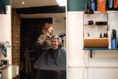 Barber styling clients hair with blow dryer in modern salon with white shelving and brick wall